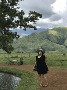 a woman in a black dress and hat stands in a grassy field with mountains in the background