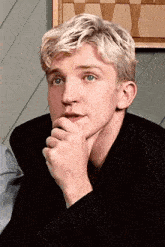 a man with blonde hair is sitting down with his hand on his chin .