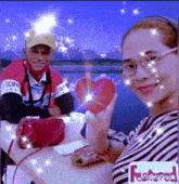 a man and a woman are sitting at a table holding a red heart in front of a body of water