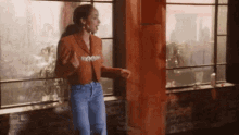 a woman in a brown jacket and jeans is dancing in front of a window
