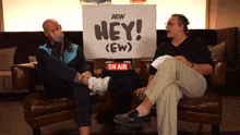 two men sit on a couch with a sign that says hey ( ew )