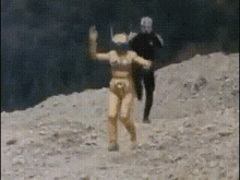 a naked woman is dancing in the dirt with a man in a black suit behind her