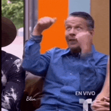 a man in a blue shirt is sitting on a couch and making a funny face with his fist in the air .