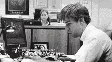 a man is sitting at a desk in front of a computer and a woman behind him .