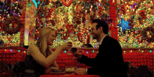 a man and a woman are sitting at a table drinking wine in front of a christmas tree