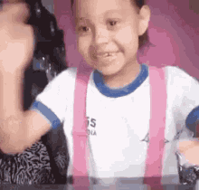 a little girl in a white shirt and pink suspenders is sitting at a table and smiling .