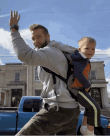 a man is carrying a child on his back in front of a blue truck with the word v8 on the back