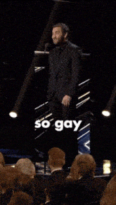 a man in a suit and tie stands in front of a microphone with the word so gay behind him
