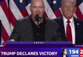 a man speaking into a microphone with the words trump declares victory on the screen behind him