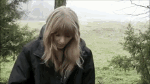 a woman wearing a black jacket is standing in a field with trees in the background .