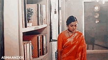 a woman in an orange saree is standing in front of a bookcase .