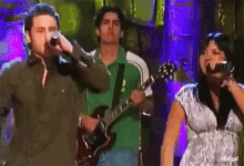 a man singing into a microphone while a man plays a guitar and a woman sings