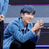 a young man with blue hair is kneeling down and clapping his hands while wearing a blue jacket .