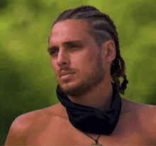 a shirtless man with braided hair and a bandana around his neck looks at the camera