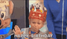 a little boy wearing a crown is standing in front of a birthday cake and says happy birthday .