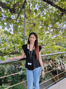 a woman in a black top and blue jeans stands in front of a lush green forest
