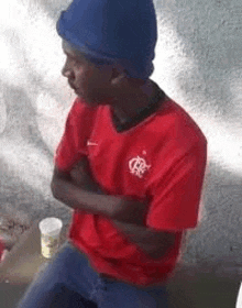 a man in a red shirt and blue hat is sitting on a bench .