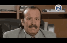 a man with a mustache is sitting in a chair in front of a shelf with the number 2 on it .