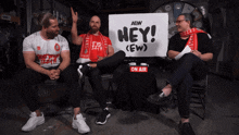 three men sitting in front of a sign that says hey