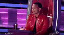 a man in a red leather jacket is sitting in a chair with a nbc logo behind him