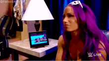 a woman with purple hair is sitting in front of a lamp watching a video on a tablet .