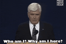 a man in a suit and tie stands at a podium and says who am i ? why am i here ?