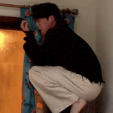 a man in a black sweater and white pants is squatting down in front of a window .