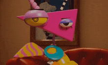 a pink triangle with a yellow eye is sitting on a red couch in front of a picture