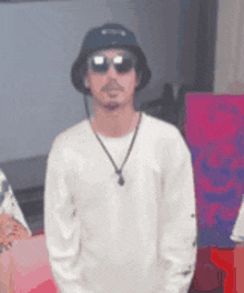 a man wearing a hat , sunglasses and a necklace is standing in front of a pink wall .