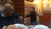 two men are sitting at a table in front of a brick wall and a sign that says ' koksal gif '