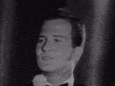 a black and white photo of a man in a tuxedo speaking into a microphone