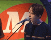 a man singing into a microphone in front of a large m on a green background