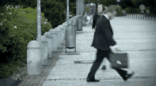 a man in a suit is walking down a sidewalk with a green briefcase