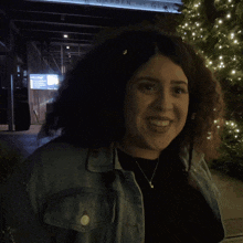 a woman wearing a denim jacket and a necklace smiles in front of a sign that says welcome