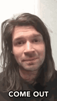 a man with long hair and a beard is smiling and saying come out .