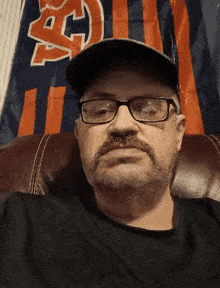 a man wearing glasses and a hat is sitting in front of a flag with the letter h on it
