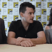 a man sits at a table in front of a wall that says comic con