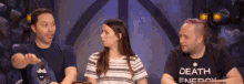 two men and a woman are sitting at a table with a woman wearing a death energy shirt .