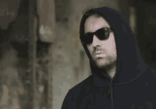 a man wearing sunglasses and a black hoodie looks at the camera