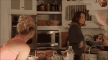 two women are standing in a kitchen talking to each other . one of the women is wearing a pink tank top .
