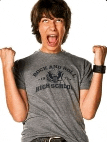 a young man wearing a gray rock and roll high school t-shirt is raising his fist in the air .
