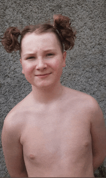a shirtless young boy with pigtails on his head looks at the camera