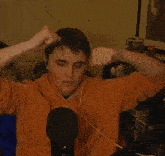 a man in an orange shirt is sitting in front of a microphone with his hands on his face