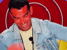 a man wearing a denim jacket and a microphone smiles in front of a red background