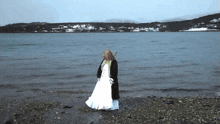 a woman in a white dress and black coat stands on a beach near a body of water