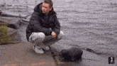 a man in a black jacket is squatting on the shore of a lake