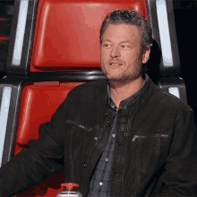 a man in a black jacket is sitting in a red chair with a red button .