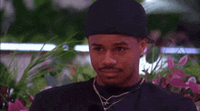 a man wearing a black hat and a necklace is talking to a woman in front of flowers .
