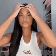a woman with long black hair is wearing a white tank top and holding her hair .