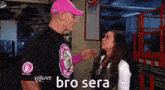 a man in a pink hat talks to a woman in a black shirt that says w live bro sera